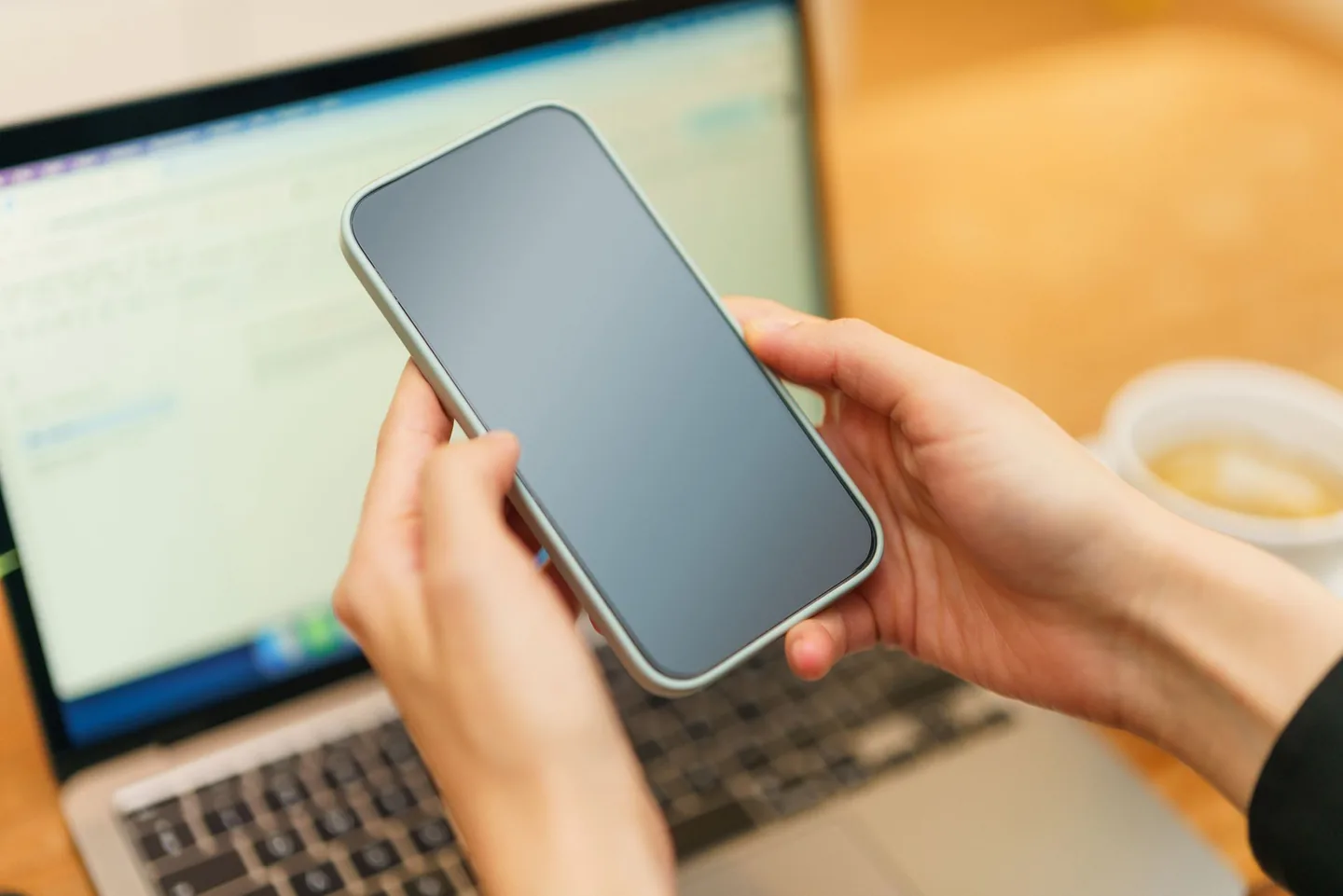 Hands holding a phone above a laptop, with a cup nearby.